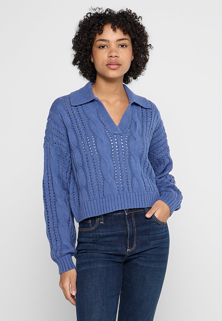 Maglione blu a maglia con collo a V e collare, caratterizzato da motivi a maglia aperta e polsini a costine, indossato con jeans in denim scuri.