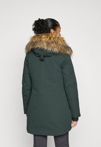 Veste rembourrée verte avec une capuche en fausse fourrure. Le manteau a une silhouette ajustée et atteint mi-cuisse, avec une finition mate et un cordon de serrage réglable.