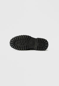 Black rubber sole with deep treads and ridges for traction, featuring a rounded toe and flat heel, marked with brand logo and design details.