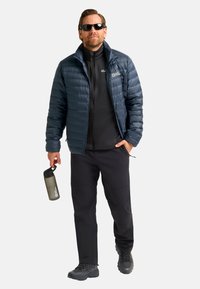 Doudoune navy avec des coutures horizontales, couche noire zippée en dessous, pantalon noir et chaussures de randonnée grises. Tiens une bouteille d'eau grise.