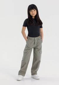 Top negro de manga corta combinado con pantalones cargo verde claro que cuentan con bolsillos laterales. La modelo lleva zapatillas blancas y está de pie con las manos en los bolsillos.