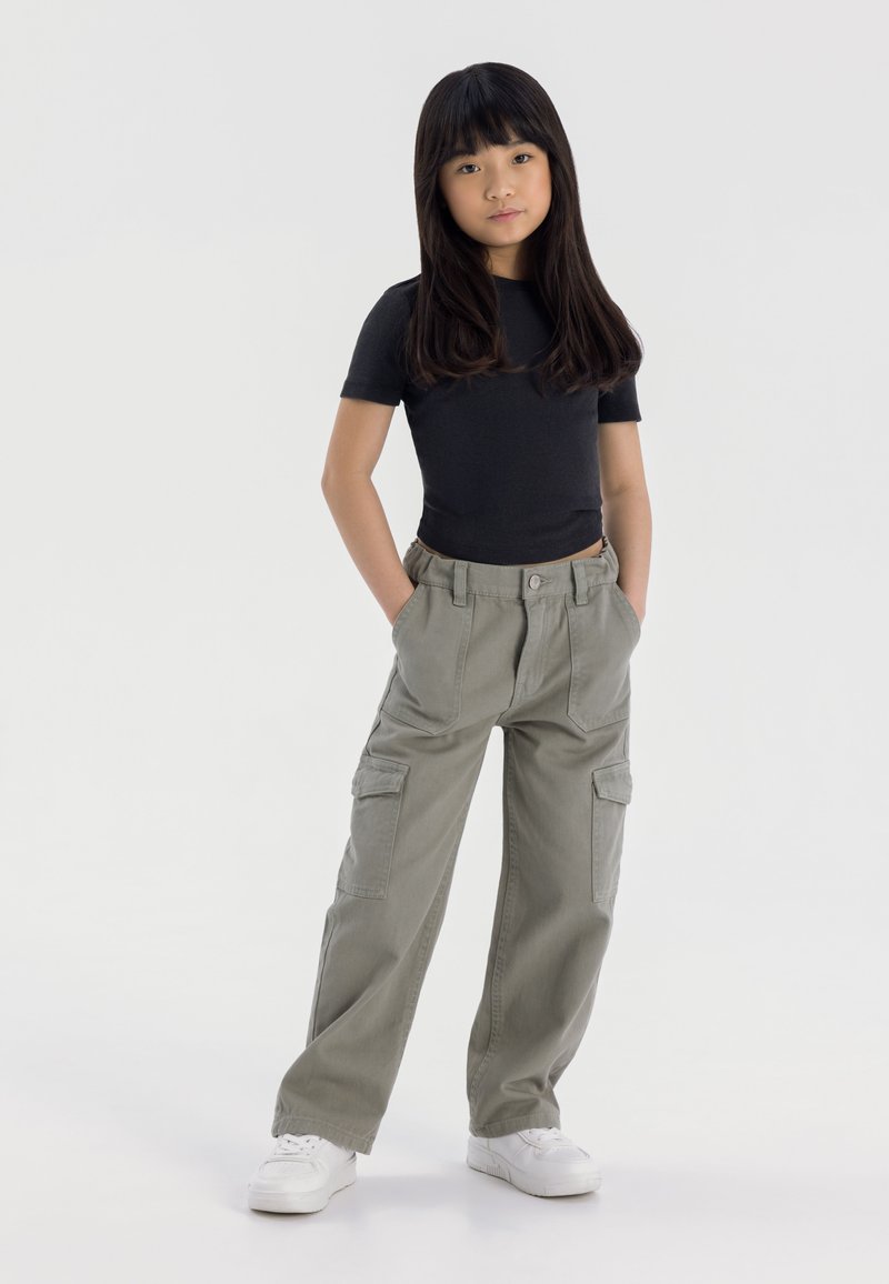 Top negro de manga corta combinado con pantalones cargo verde claro que cuentan con bolsillos laterales. La modelo lleva zapatillas blancas y está de pie con las manos en los bolsillos.