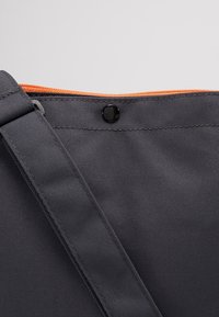 Šedá látková taška s oranžovým zipem, vybavená černým patentním knoflíkem, texturovaným povrchem a širokým nastavitelným popruhem.