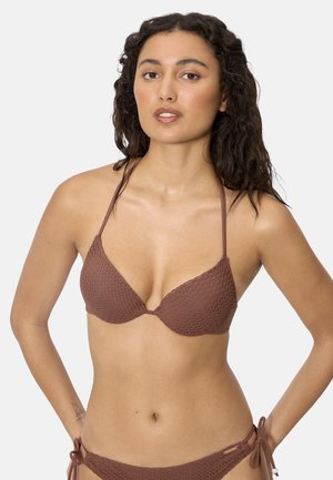 Mujer con cabello oscuro y ondulado, vistiendo un top de bikini marrón texturizado de tipo halter y braguitas a juego con lazos, sobre un fondo liso.