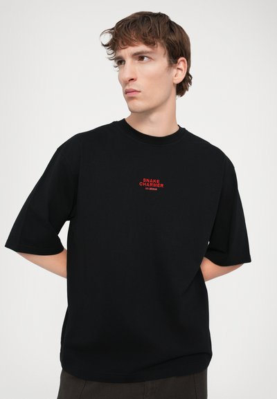 T-shirt en coton noir avec col rond, manches courtes et inscription rouge "SNAKE CHARMER" centrée sur la poitrine, offrant une coupe décontractée.