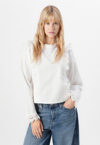Giovane donna con capelli biondi indossa una blusa bianca con pizzo e jeans blu, posando con una mano in tasca su uno sfondo bianco.
