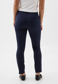 Tamsiai mėlyni aptempti kelnės su lygiu audiniu, dviem kišenėmis gale ir siaurėjančiomis kojų apačiomis, derinamos su juodais lygiais batais.