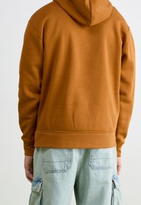 Persona vestida con una sudadera de color óxido y pantalones cargo de mezclilla azul claro, de espaldas a la cámara contra un fondo simple.