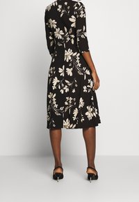 Czarna sukienka w kwiaty z trzema ćwierć rękawami, ozdobiona białymi kwiatami i zielonymi akcentami. Sukienka ma rozkloszowany dół i gładką teksturę.