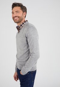 Maglione grigio a collo a V realizzato in materiale morbido, indossato sopra una camicia a quadri marrone e blu con bottoni. Abbinato a jeans in denim scuro.