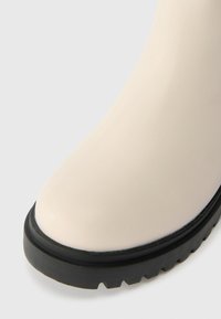 Beige vedenpitävät saappaat, joissa on sileä pinta, pyöreä kärki ja musta kumipohja, jossa on syvä uritus pitävyyttä varten.