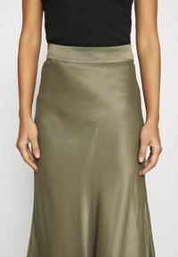 Buste féminin portant un haut noir sans manches et une jupe midi en satin vert olive, mains détendues le long du corps sur fond blanc.