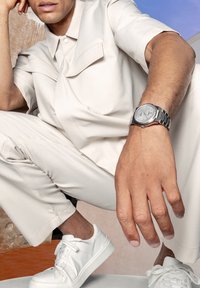 Chemise blanche ajustée avec poches avant, pantalon slim assorti, montre en argent avec un cadran rond et baskets blanches. Texture de tissu lisse.