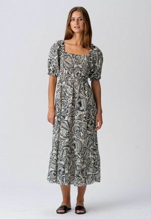 Une femme porte une robe midi noire et blanche à motifs avec des manches bouffantes et des sandales plates noires, posant devant un fond clair uni.