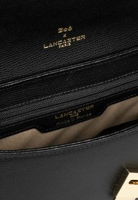 Intérieur d'un sac en cuir noir avec une fermeture éclair dorée et des étiquettes de marque en relief, doublé d'une douce teinte tan, de forme structurée et au design minimaliste.