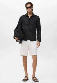 Sort button-up skjorte, hvide shorts og sorte sandaler. Skjorten har en afslappet pasform og to forlommer. Modellen holder en sort taske.