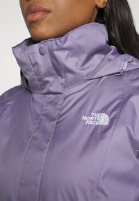 Gros plan d'une personne portant une veste North Face violette avec un col montant et une fermeture à boutons-pression.