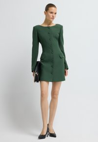 Frau in einem figurbetonten, langärmeligen grünen Kleid mit Knöpfen, die eine schwarze Clutch-Tasche hält, schwarze High Heels tragend, vor einem weißen Hintergrund stehend.