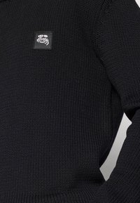 Black knitted sweater featuring a textured fabric. A small square logo tag with white design is positioned on the front upper left.