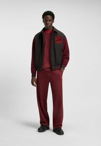 Maronfarvet træningssæt bestående af en pullover og bukser med ribbet manchetter. Accentueret med en sort varsity-jakke og rød broderi. Sorte sko.