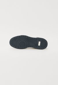 Suela de zapatilla de goma negra con patrones texturizados y ranuras. Presenta un acento rectangular blanco cerca del talón. Perfil plano para mayor estabilidad.