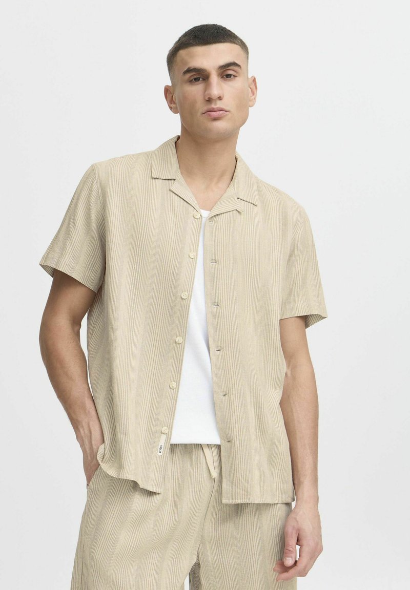 Camisa de manga corta a rayas beige, hecha de tejido suave, con cuello tipo notch y cierre de botones en la parte delantera, combinada con pantalones cortos claros a juego.