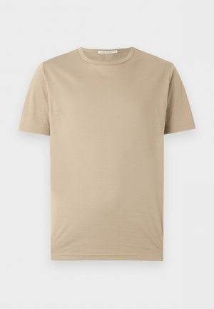 Jeune homme debout, main dans la poche, portant un t-shirt beige à manches courtes, un jean large bleu clair et des chaussures noires sur fond blanc.