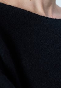 Primo piano del collo e della spalla di una persona che indossa un maglione nero texturizzato con un'ampia scollatura.