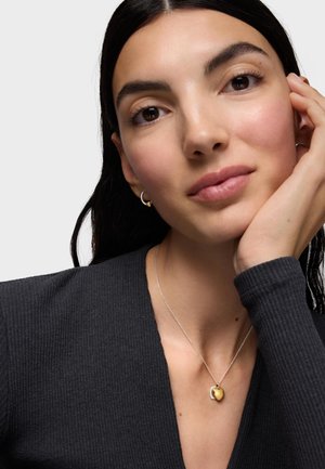 Donna con capelli scuri che indossa piccoli orecchini a cerchio e un ciondolo a forma di cuore in oro su una catena d'argento, appoggiando il viso sulla mano.