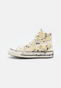 Sneaker montante blanche avec un design éclaboussé de peinture noire et jaune, lacets blancs et semelle en caoutchouc avec des détails grisés noirs.
