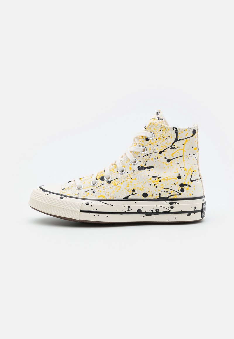 Sneaker montante blanche avec un design éclaboussé de peinture noire et jaune, lacets blancs et semelle en caoutchouc avec des détails grisés noirs.