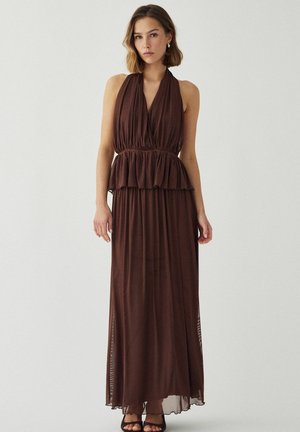 Frau trägt ein ärmelloses, tiefbraunes Neckholderkleid mit taillierter Taille, Peplum-Detail und einem fließenden langen Rock, steht vor einfachem Hintergrund.