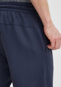 Sötétkék sportos jogging nadrág elasztikus derékkal, oldal zsebekkel és sima felülettel. Az anyag enyhe fényes hatású, és a szegélyeknél szűkített.