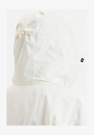 Biała kurtka przeciwdeszczowa z kapturem, o gładkiej fakturze, z bocznym sznurkiem do regulacji oraz małą czarną metką na plecach.