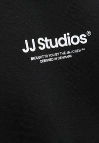 Black cotton garment featuring white text: "JJ Studios," with additional phrases below. Text is clean and clearly laid out.