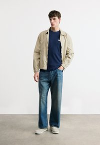 Νέος άνδρας που στέκεται με το χέρι στην τσέπη, φοράει μπεζ σακάκι, μπλε σκούρο πουκάμισο, φαρδιά μπλε τζιν και λευκά αθλητικά παπούτσια πάνω σε γκρι πάτωμα.