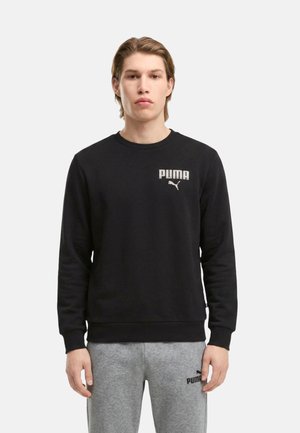 Joven con una sudadera negra de Puma y pantalones deportivos grises de Puma de pie frente a un fondo claro y liso.