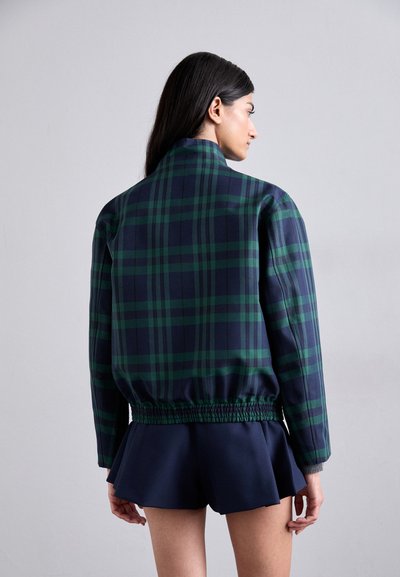 Giacca a quadri in blu navy e verde con vestibilità rilassata, fascia elastica in vita e colletto erettore. Indossata sopra una gonna plissettata blu navy.