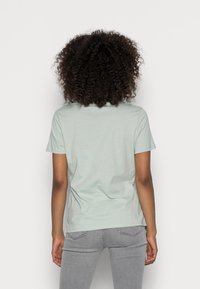 Mujer con cabello rizado que lleva una camiseta de mangas cortas de color verde claro y jeans grises, de espaldas frente a un fondo blanco liso.