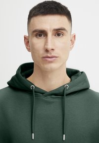 Sudadera verde con cordón, con ojales plateados. Textura suave con un ajuste relajado, que muestra un diseño minimalista.