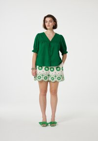 Groene linnen knooptop met pofmouwen, gecombineerd met witte shorts met geschulpte rand en groene bloemenborduursels. Groene strik sandalen maken de look compleet.