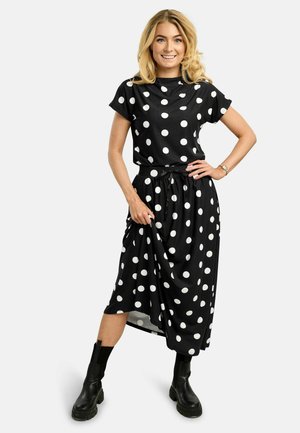 Lyshåret kvinde iført en sort kjole med store hvide polkaprikker, sorte ankelstøvler, smilende med den ene hånd på hoften og den anden hånd, der holder kjolen.