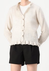 Beige stickad cardigan med vågig nederkant, knäppning fram och långa ärmar, tillsammans med svarta shorts. Mjuk textur och avslappnad design.