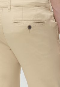 Gros plan sur un pantalon beige montrant une poche arrière passepoilée avec un bouton foncé et une boucle de ceinture à la taille et à la hanche d’une personne.