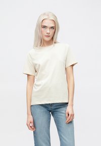 Beidža īsām piedurknēm T-krekls, kas izgatavots no kokvilnas. Klasiska, apaļa kakla izgriezuma, taisna piegriezuma, gluda tekstūra un minimālas detaļas. Valkāts ar gaiši ziliem džinsiem.