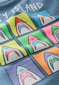 Camiseta de color azul claro con un diseño de cuadrícula que presenta caras de tiburones en estilo de dibujos animados en varios colores: rosa, verde, naranja y azul, con contornos gruesos y detalles en los dientes.