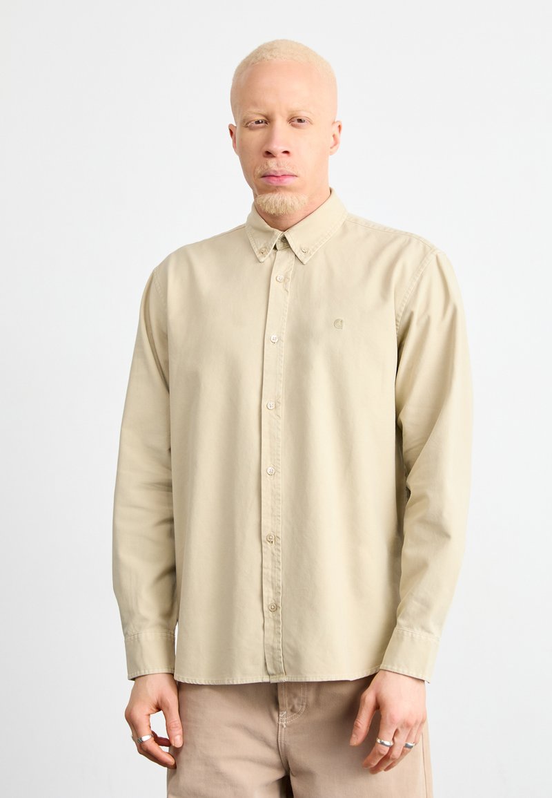 Chemise à manches longues beige à boutons avec col, en tissu lisse ; présente un logo discret sur la poitrine et une coupe décontractée.