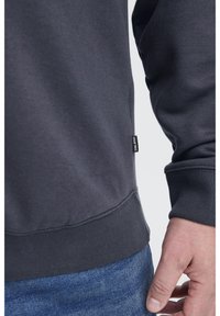 Közelkép egy sötétszürke pulóveres személyről, aki "BLEND" felirattal rendelkezik és kék farmert visel, a könyök és a kéz egy része lazán a teste mellett.