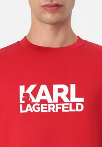 Camiseta de algodón roja de cuello redondo con un destacado logo blanco que presenta un diseño de silueta, textura suave y detalle de cuello acanalado.