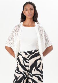 Cardigan en maille en dentelle de couleur blanc cassé, avec une coupe ample et des manches courtes. Associé à un pantalon noir et beige à motifs.